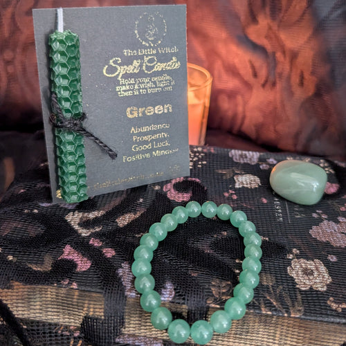 A green hand rolled beeswax candle mounted onto black card with gold foil The Little Witch branding, a green aventurine tumbled crystal and a green aventurine crystal bracelet all set upon a black floral lace background