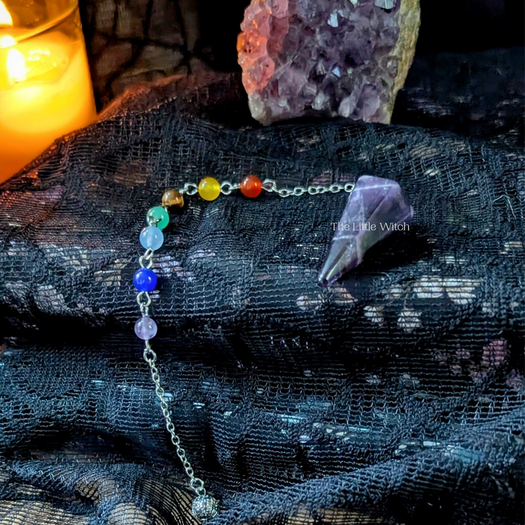 Amethyst pendulum with chakra crystal beads on a textured surface with crystals and a candle in the background