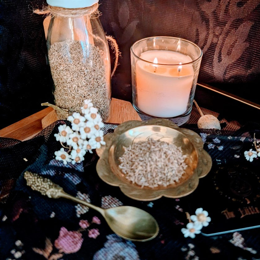 Candle, jar, and bowl of aniseeds on a decorative tablecloth with flowers and a spoon.