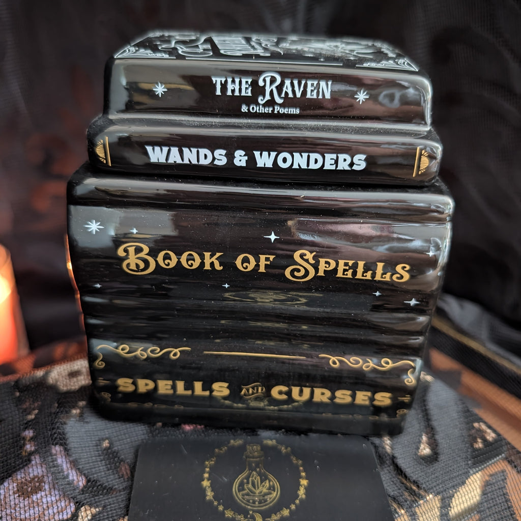 A stack of four glossy black book-shaped items with decorative gold and white text reading 'The Raven & Other Poems,' 'Wands & Wonders,' 'Book of Spells,' and 'Spells & Curses,' photographed against a dark fabric background.