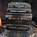 A stack of four glossy black book-shaped items with decorative gold and white text reading 'The Raven & Other Poems,' 'Wands & Wonders,' 'Book of Spells,' and 'Spells & Curses,' photographed against a dark fabric background.
