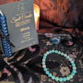 A blue beeswax candle mounted onto black card with gold foil The Little Witch branding, an amazonite crystal bracelet and a sodalite crystal upon a black floral lace background