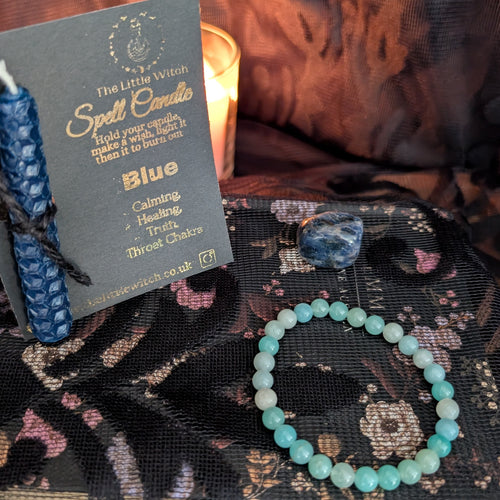 A blue beeswax candle mounted onto black card with gold foil The Little Witch branding, an amazonite crystal bracelet and a sodalite crystal upon a black floral lace background