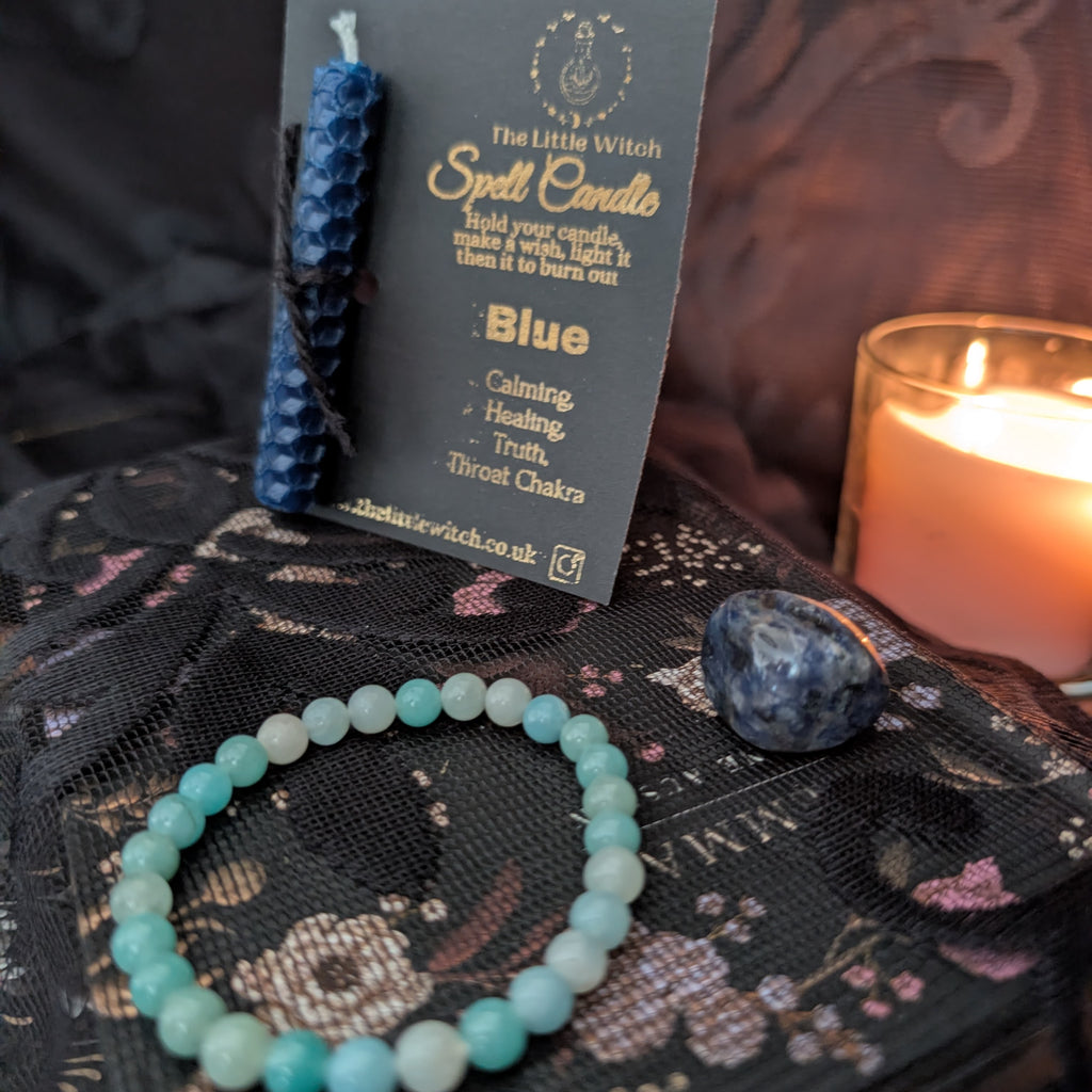 A blue beeswax candle mounted onto black card with gold foil The Little Witch branding, an amazonite crystal bracelet and a sodalite crystal upon a black floral lace background