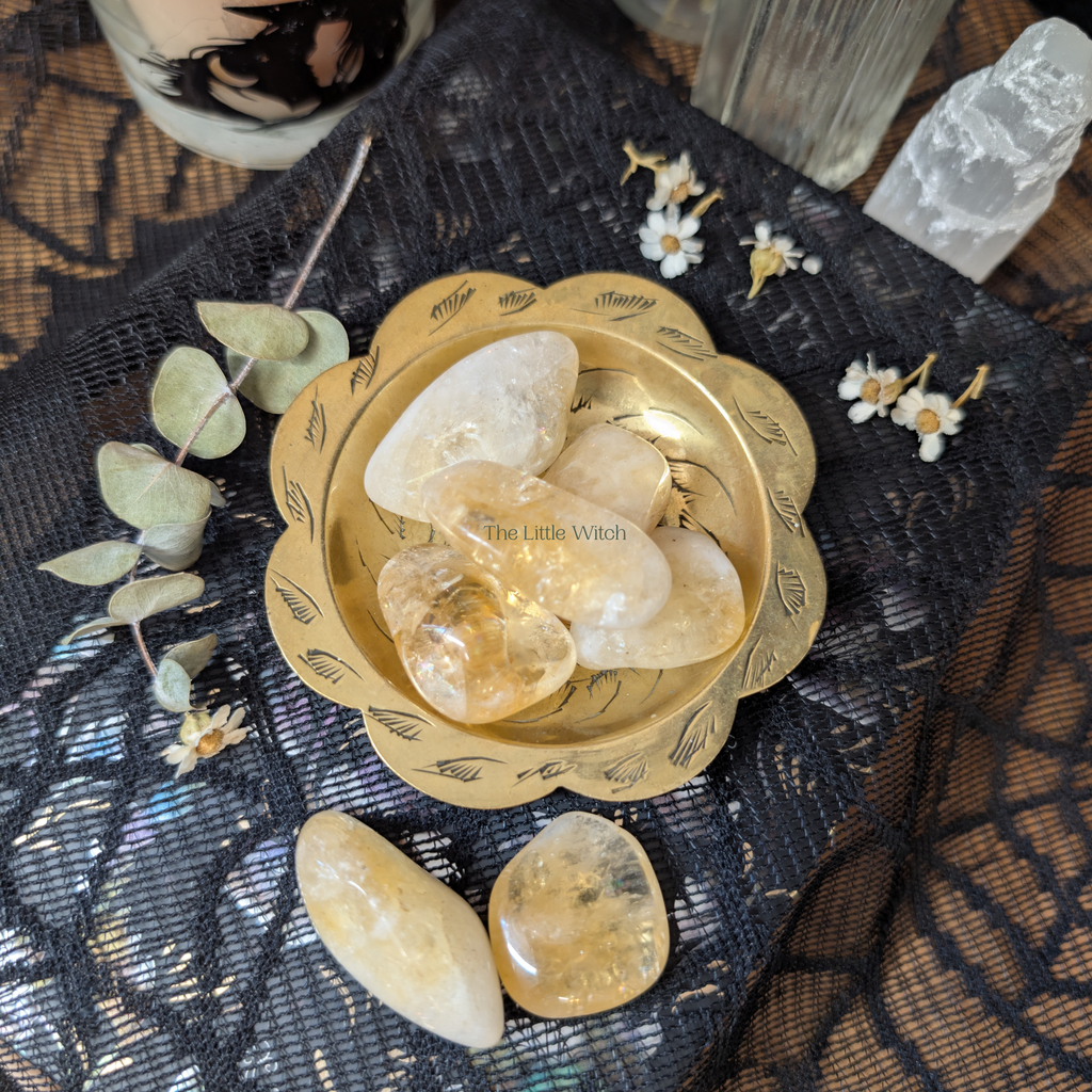 Decorative bowl with stones and flowers on a textured surface, featuring 'The Little Witch' branding.