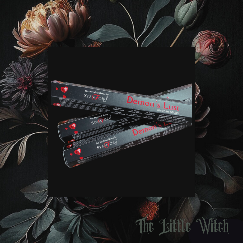 A box of Demon's Lust incense sticks placed among green leaves and flowers, with the brand name 'The Little Witch' visible.