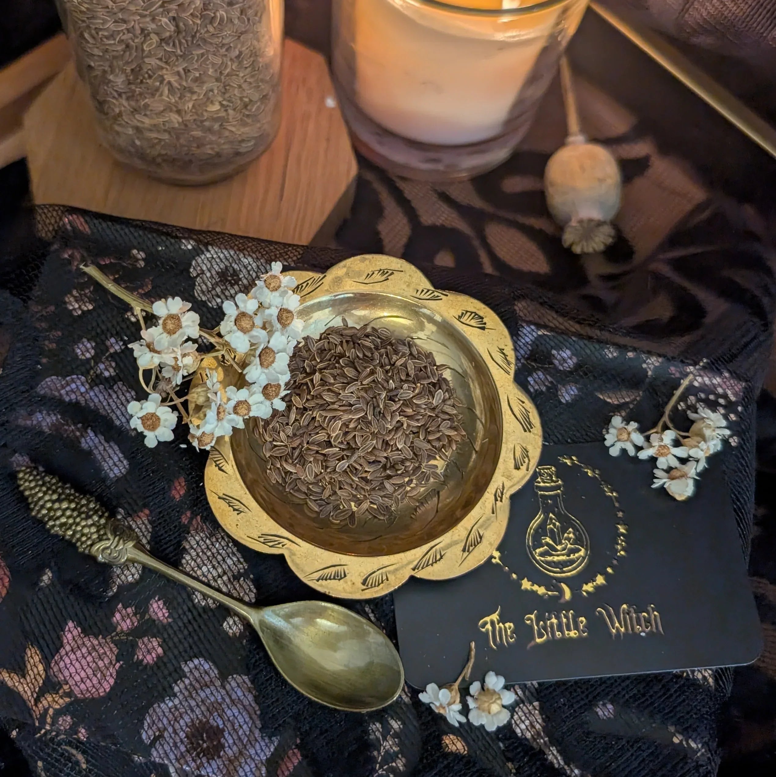 Decorative setup with a gold container of herbs, a spoon, and a card labeled 'The Little Witch' on a dark fabric background.