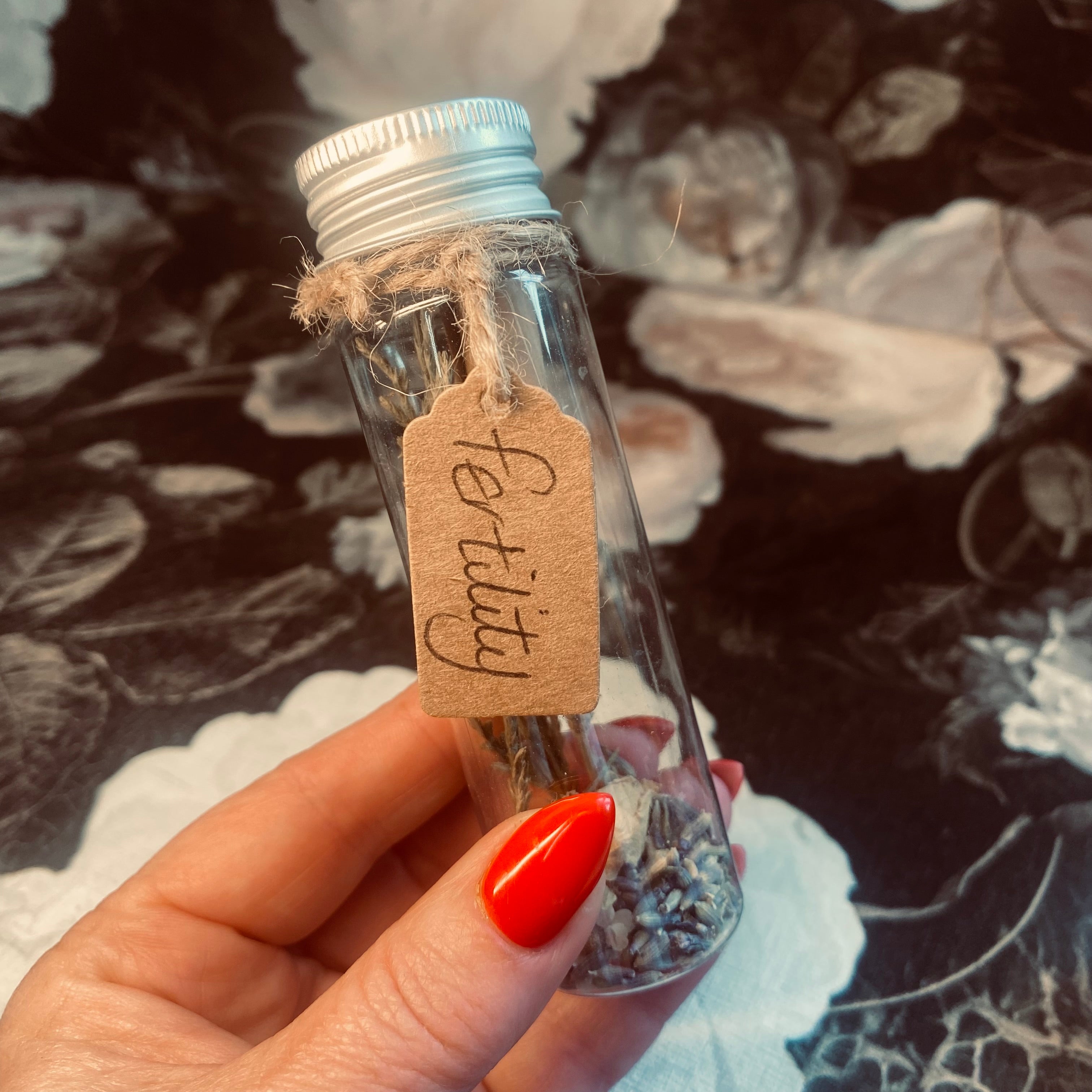 A hand holding a clear tube with a label that reads 'Fertility', containing herbs and flowers for a fertility spell. The tube is sealed with a piece of paper with an affirmation message.