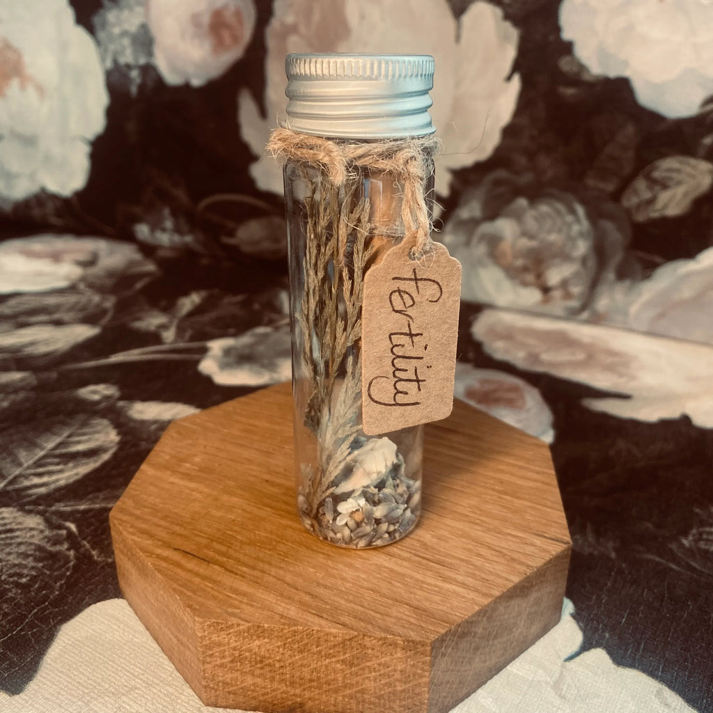 A hand holding a clear tube with a label that reads 'Fertility', containing herbs and flowers for a fertility spell. The tube is sealed with a piece of paper with an affirmation message.