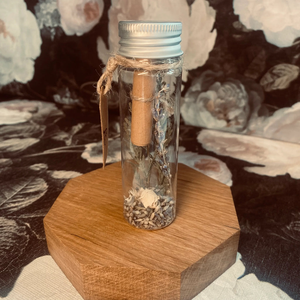 A hand holding a clear tube with a label that reads 'Fertility', containing herbs and flowers for a fertility spell. The tube is sealed with a piece of paper with an affirmation message.