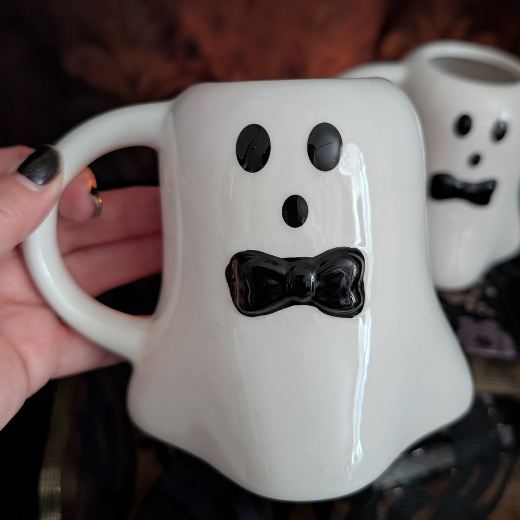 A white ceramic ghost-shaped mug with a black bow-tie face detail, held by a hand with black nail polish. A second matching ghost mug is visible in the background.