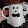 A white ceramic ghost-shaped mug with a black bow-tie face detail, held by a hand with black nail polish. A second matching ghost mug is visible in the background.