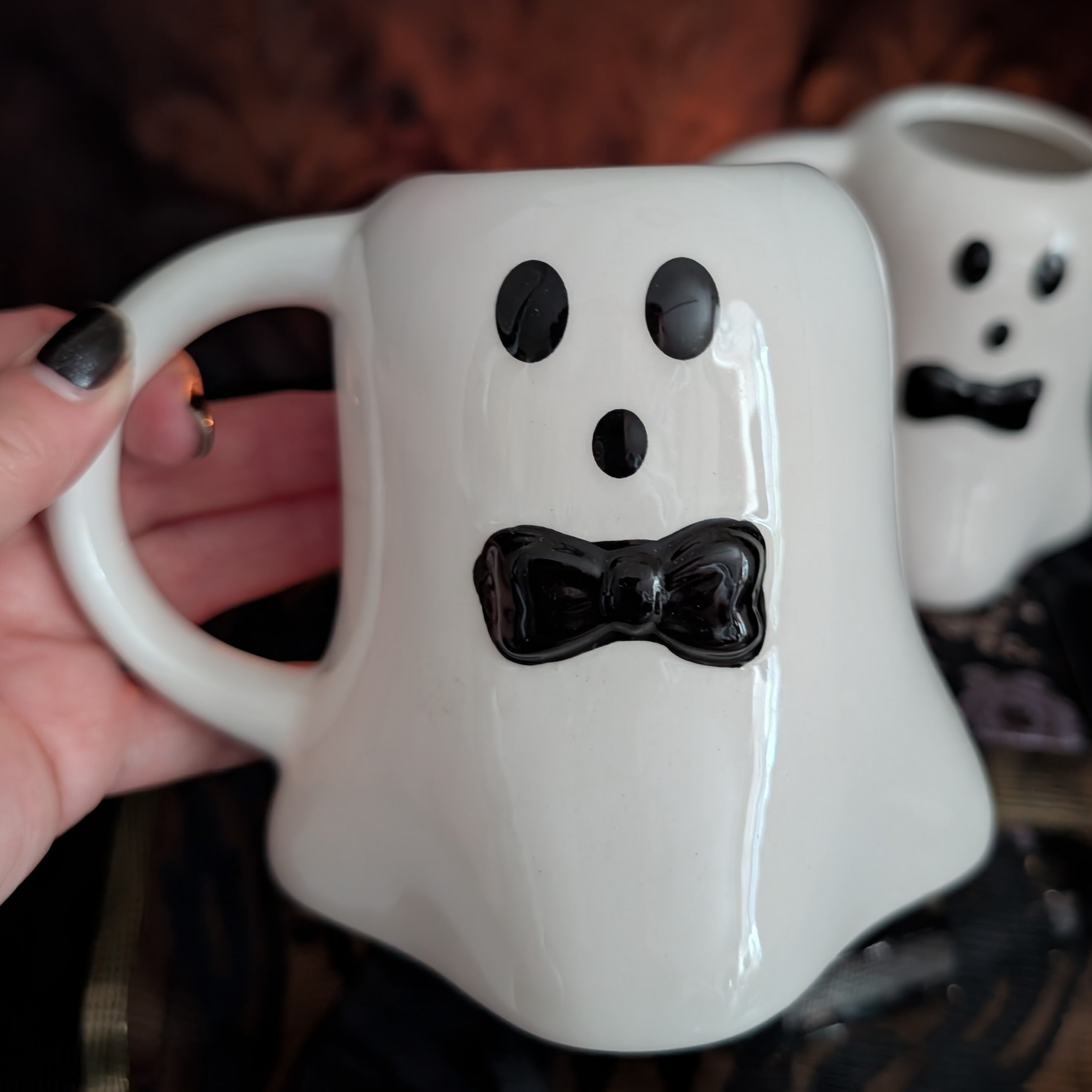 A white ceramic ghost-shaped mug with a black bow-tie face detail, held by a hand with black nail polish. A second matching ghost mug is visible in the background.