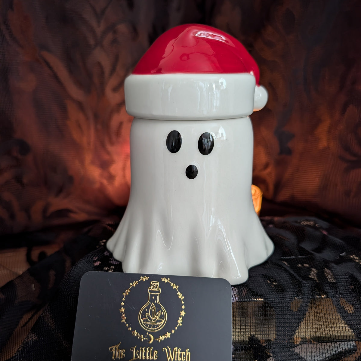 A white ceramic ghost-shaped oil burner with a red Santa hat, photographed against a dark patterned fabric background with a black lace cloth in front and a small card labeled 'The Little Witch' at the bottom.