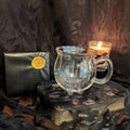 Clear glass mug on a dark surface with a candle and black pouch in the background.