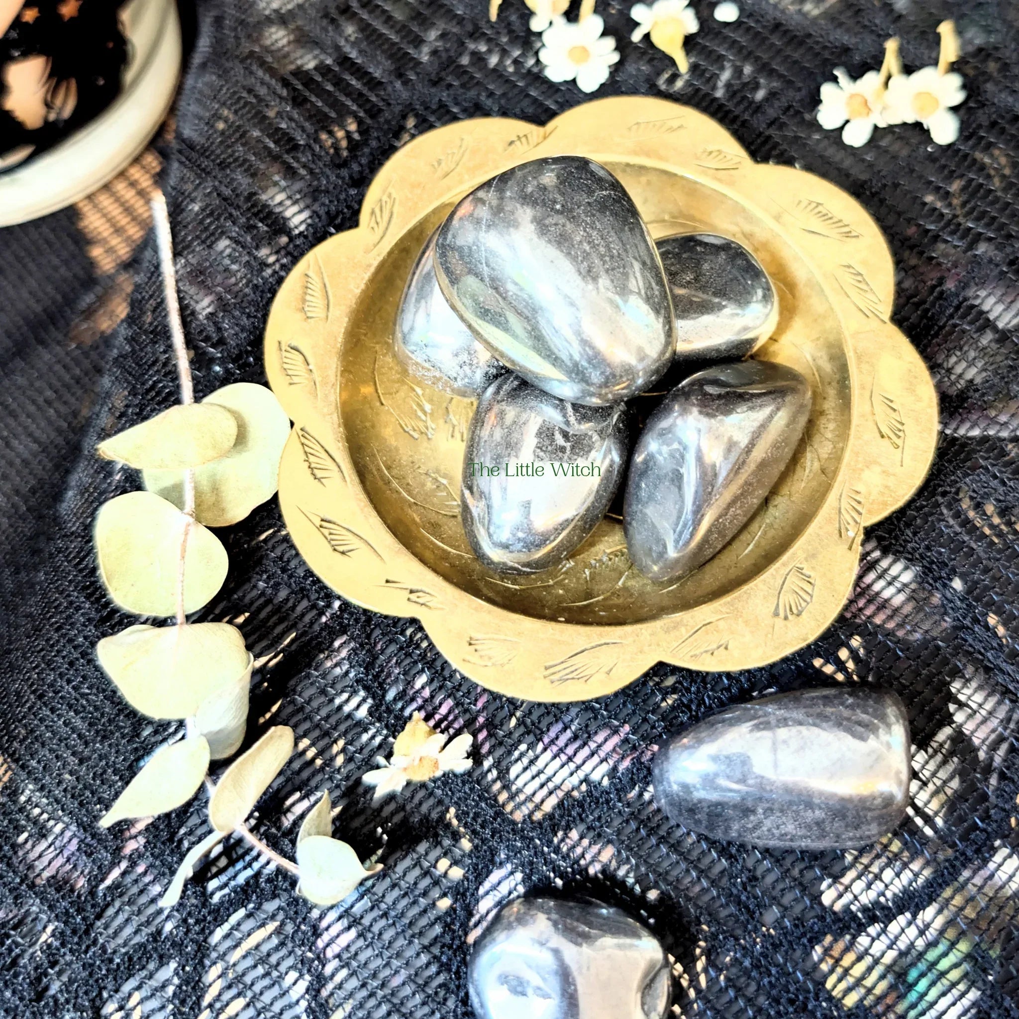A collection of Hematite crystals in a gold dish on a dark fabric background with flowers.