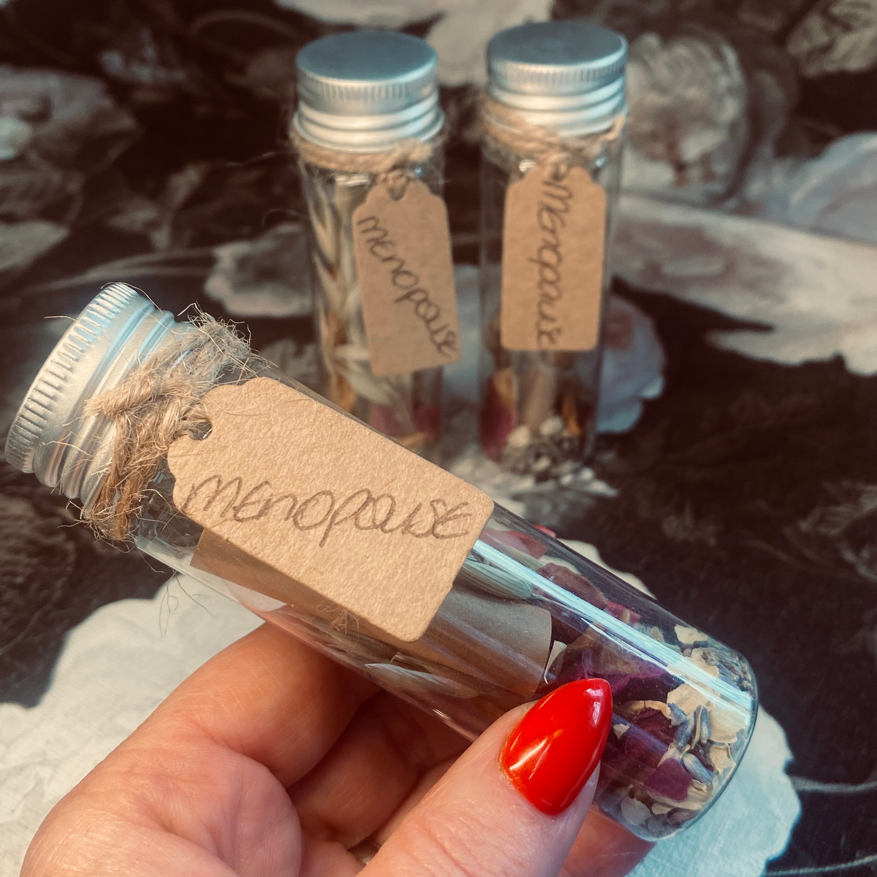 A hand holding a clear glass jar with a label that reads 'MENOPAUSE', containing herbs and flowers. Two more jars with similar labels are visible in the background.