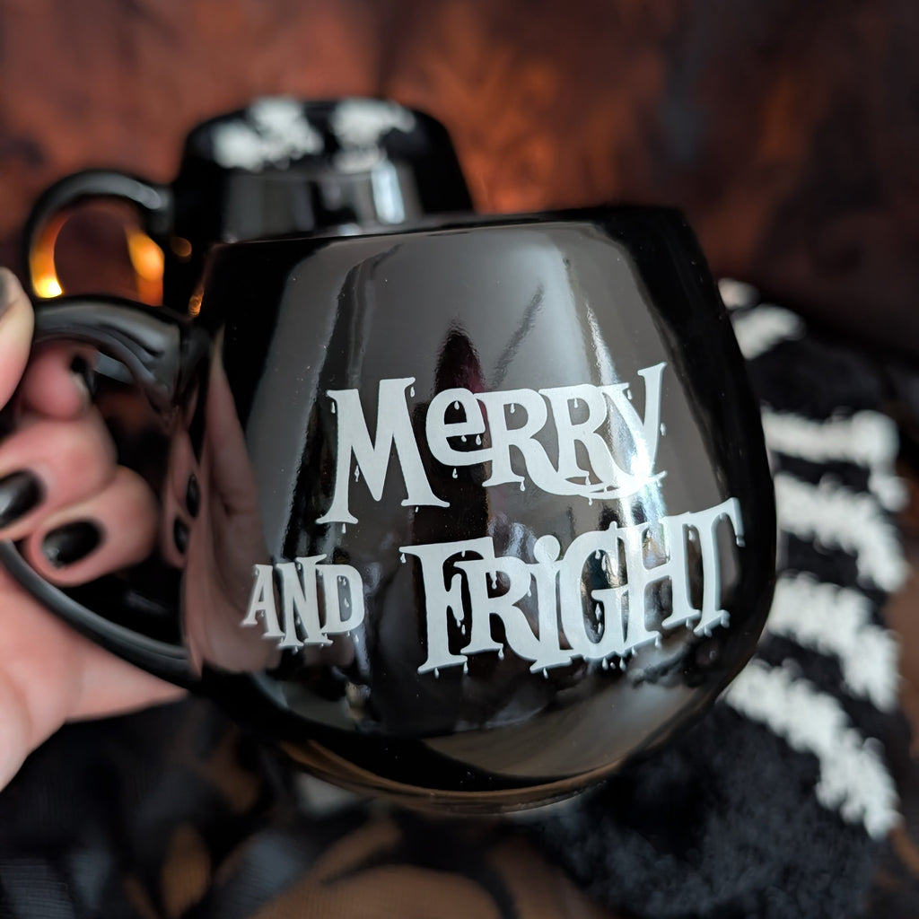 A hand with black nail polish holds a glossy black ceramic mug featuring the words 'MERRY AND FRIGHT' in a white horror-style font. A second matching mug is visible in the background against a dark, warm-toned backdrop.
