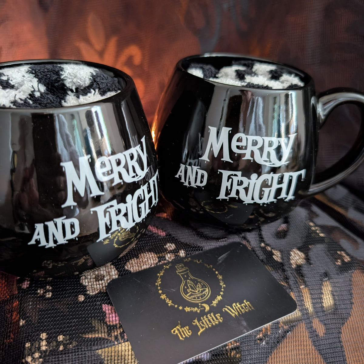 Two black ceramic mugs with white gothic text reading 'Merry and Fright' are shown on a patterned black fabric surface. A small black card with gold lettering reading 'The Little Witch' is placed in front.