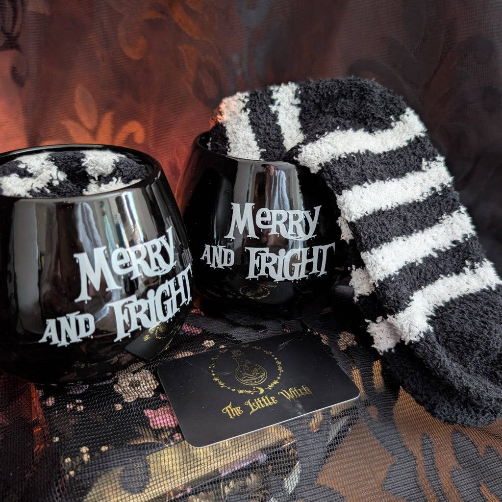A cozy Halloween-themed gift set featuring two black stemless wine glasses with white 'Merry and Fright' text, a black and white striped fluffy socks draped over one glass, and a black card reading 'The Little Witch' with a gold emblem, arranged on a dark patterned fabric surface.