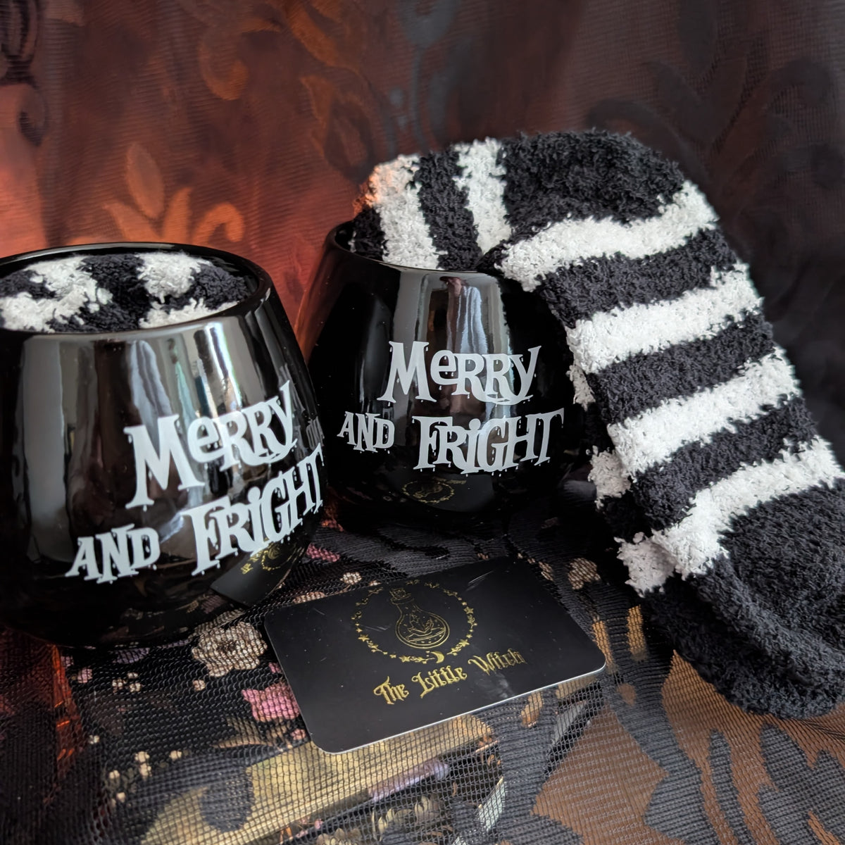 A cozy Halloween-themed gift set featuring two black stemless wine glasses with white 'Merry and Fright' text, a black and white striped fluffy socks draped over one glass, and a black card reading 'The Little Witch' with a gold emblem, arranged on a dark patterned fabric surface.