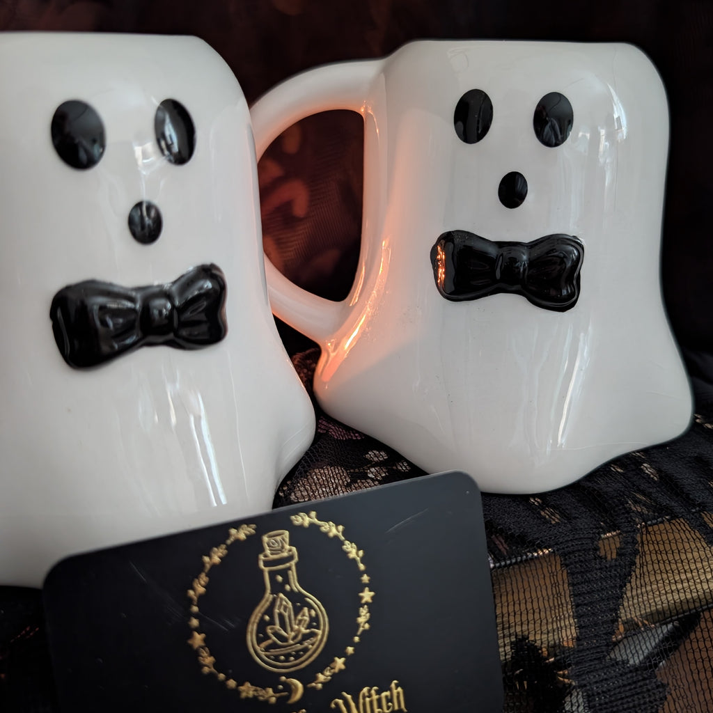 Two white ceramic mugs shaped like cartoon ghosts with black eyes, noses, and bow ties, shown on a dark lace surface with a black card featuring a gold witch potion logo in the foreground.