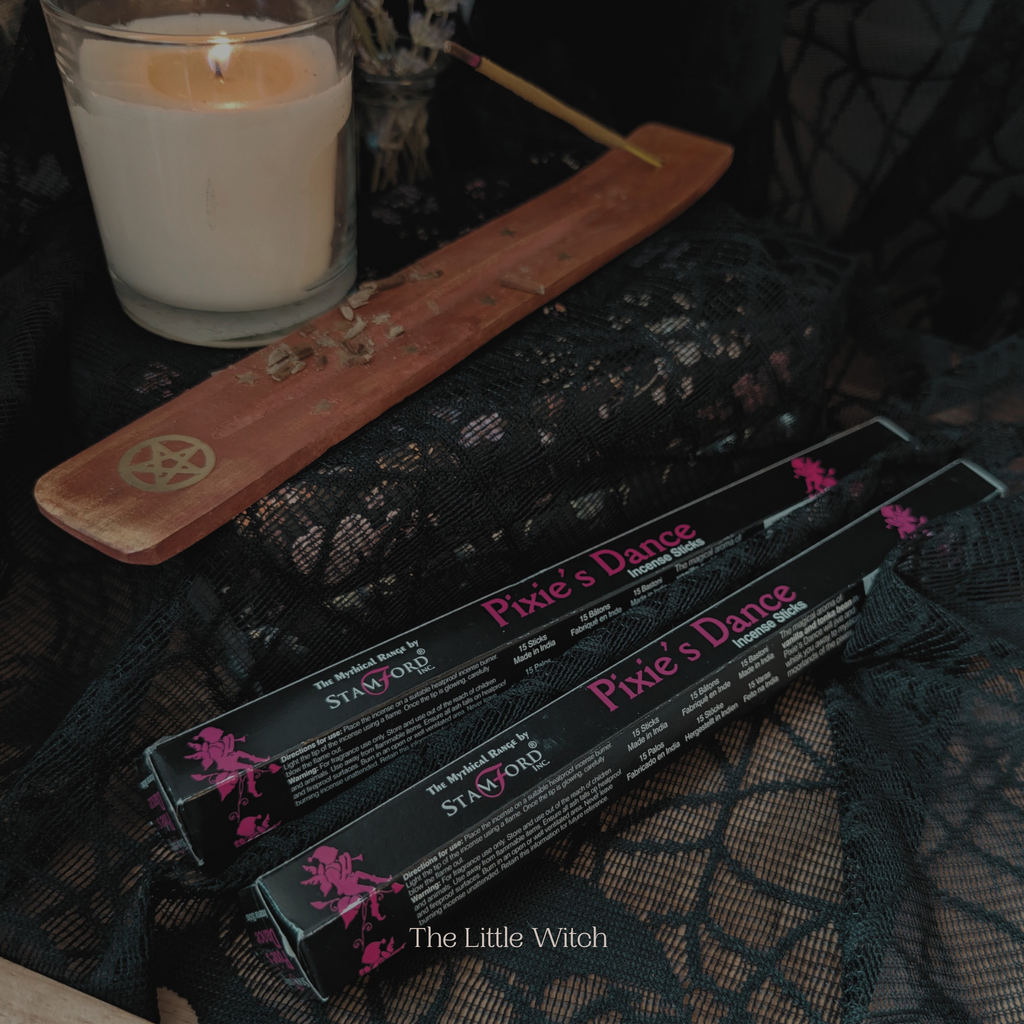 Three boxes of 'Pixie's Dance' with a lit candle and wooden pentacle on a dark fabric background.