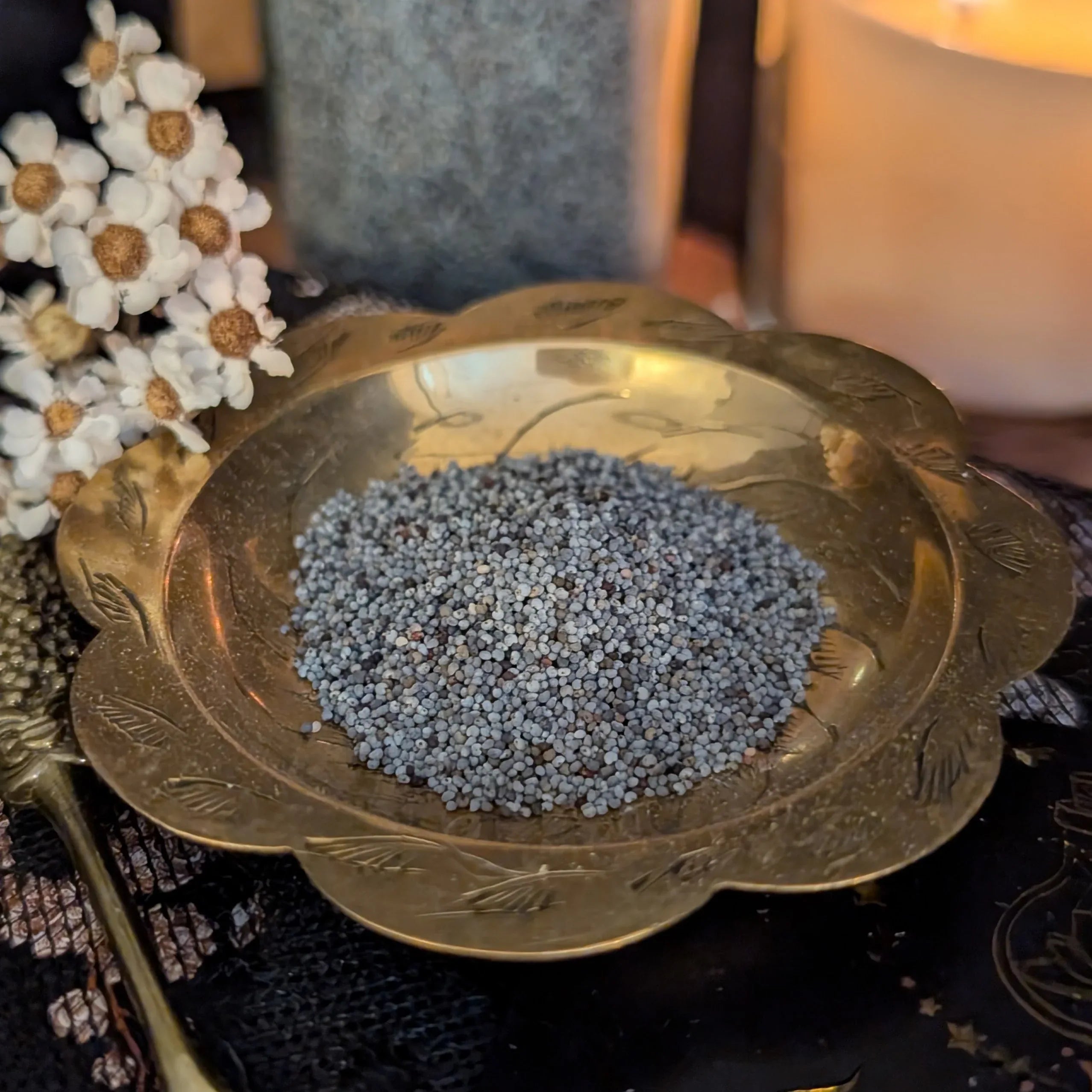 Decorative brass bowl with dried poppy seeds on a dark surface with flowers and a candle in the background.