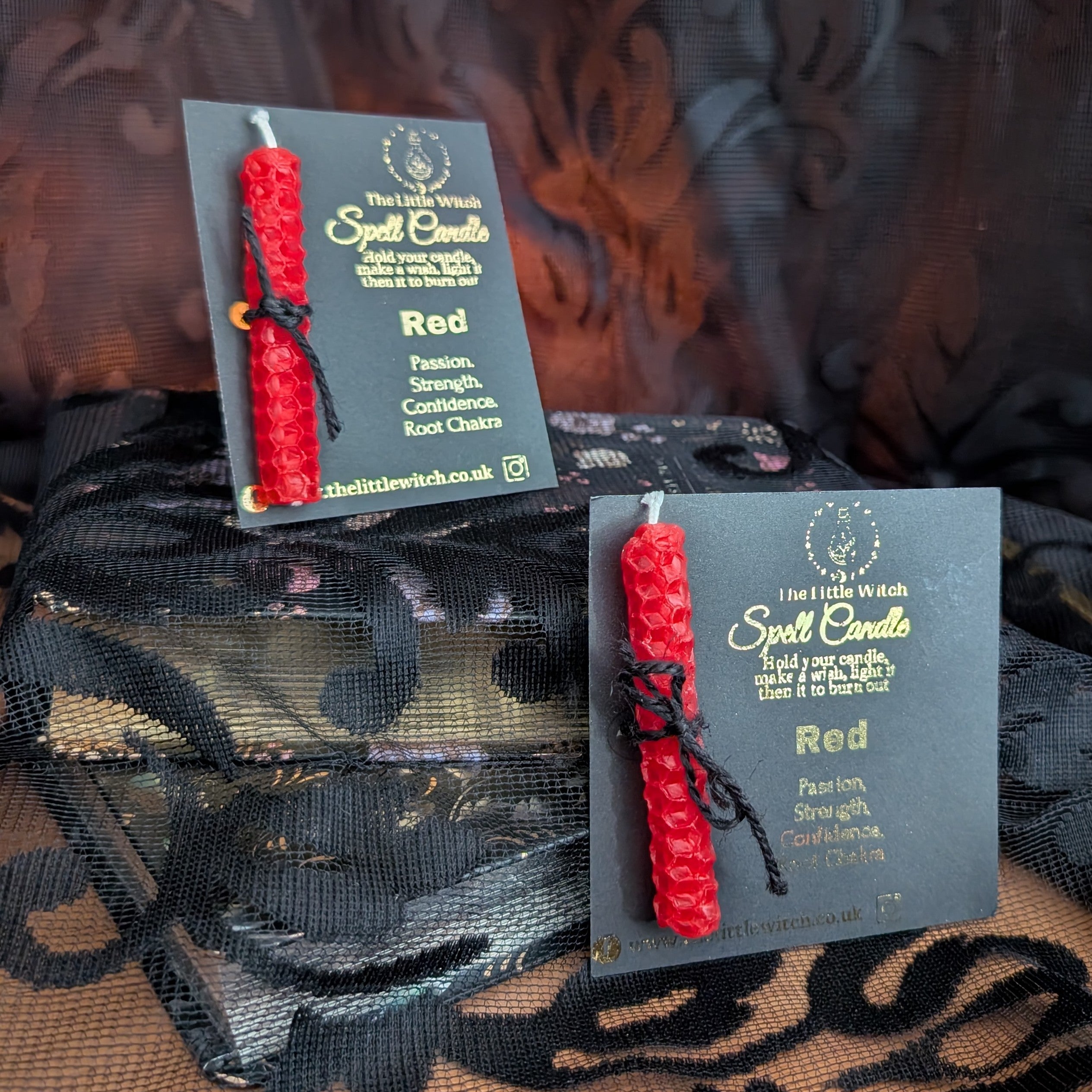 Two red spell candles with black ribbons on cards, placed on a decorative fabric background.