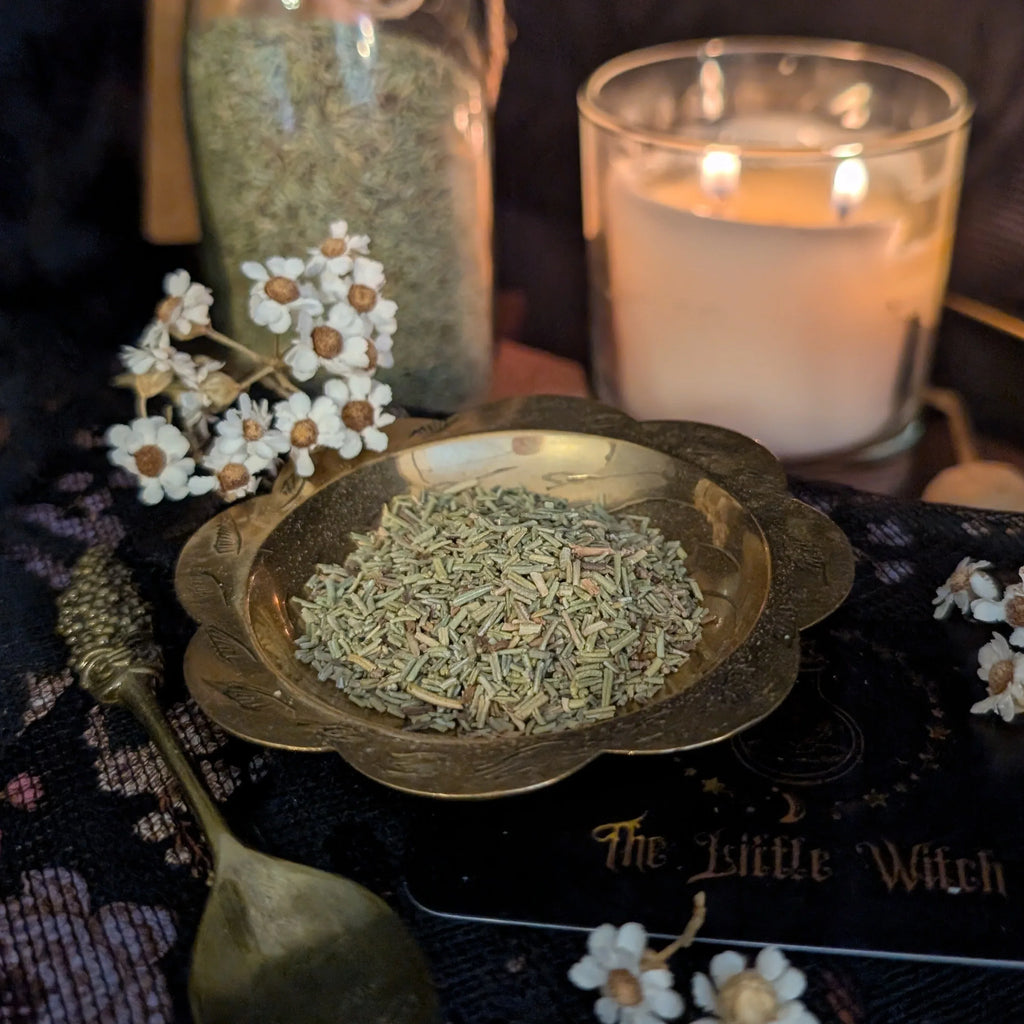 Dried Rosemary in a brass dish with a lit candle and decorative elements on a dark surface.