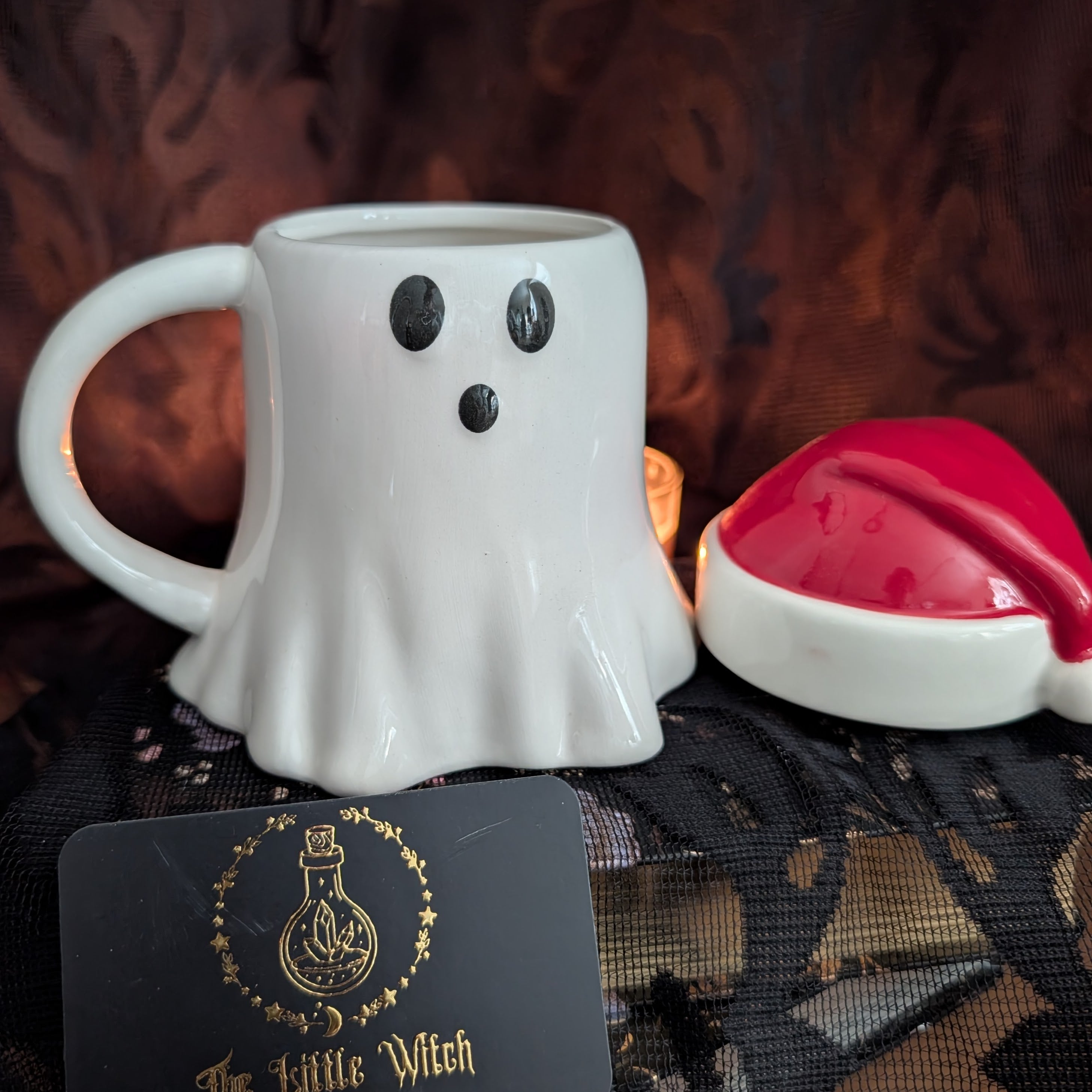 A white ceramic ghost-shaped mug with black eyes and a mouth, displayed next to a red and white Santa hat and a black card with gold lettering that reads 'The Little Witch'.