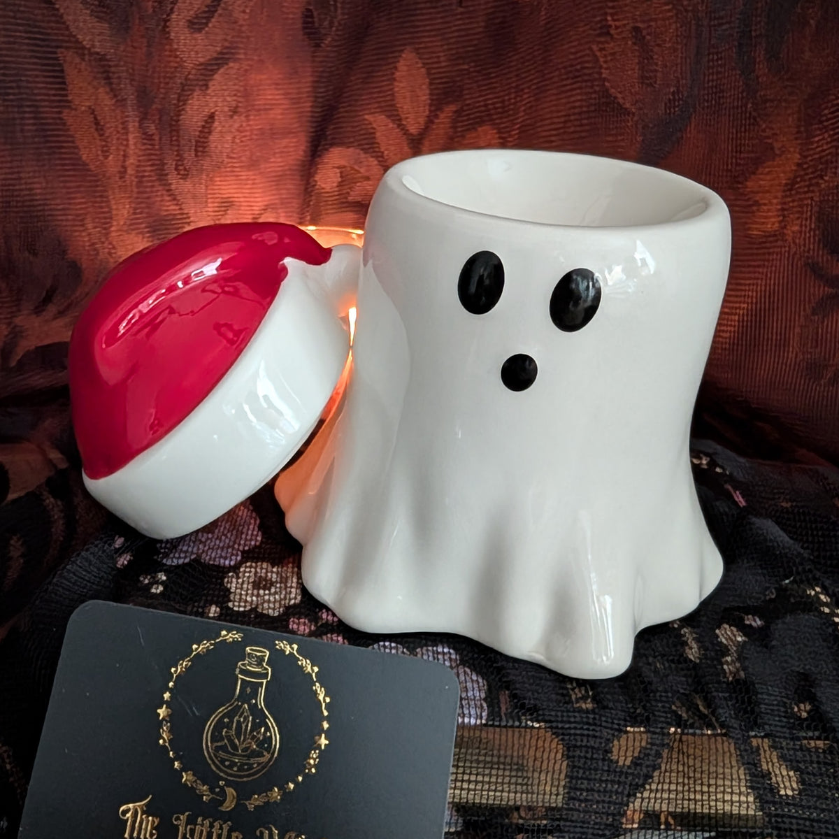 A white ceramic ghost-shaped wax warmer with black eyes and mouth, shown with its red-and-white lid leaning beside it. The warmer is placed on a dark patterned fabric surface with a warm glowing candle inside, and a small branded card is visible in the foreground.