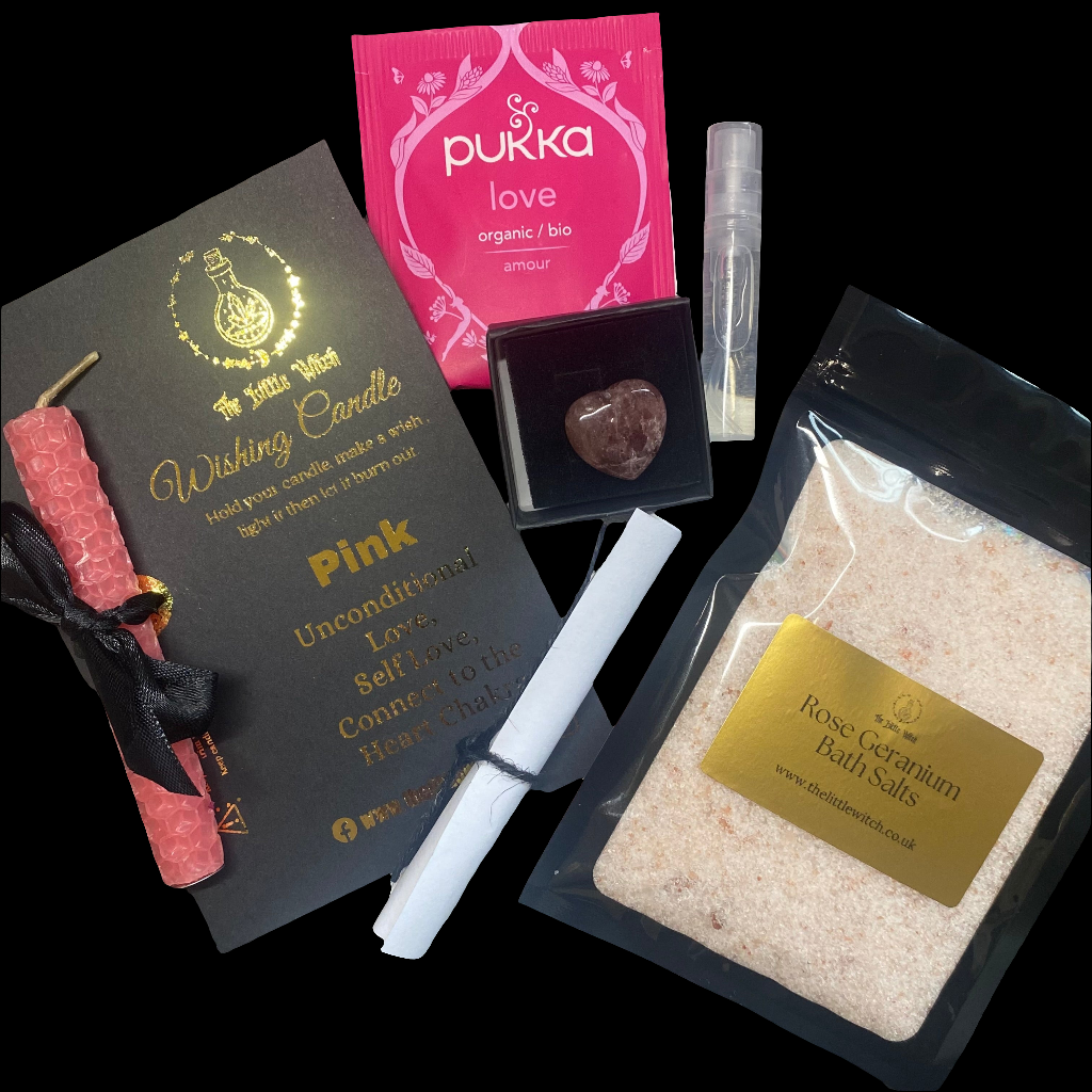 Pink beeswax candle mounted on black card, a strawberry quartz heart shaped crystal in a gift box, an organic pink sachet of 'Love' pukka tea, mini energy cleansing spray and Rose Geranium Bath salts on a black background