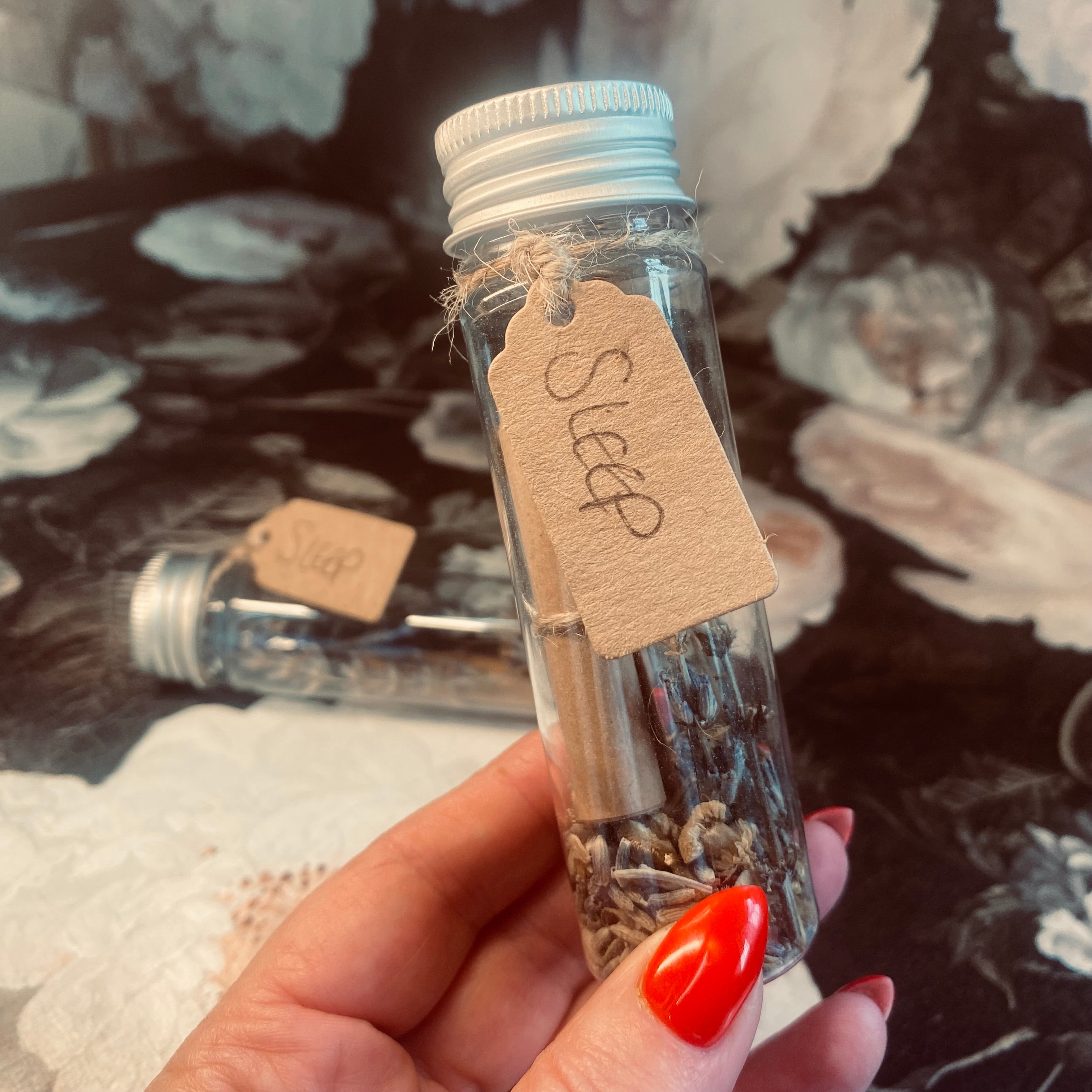 A hand holding a small clear glass jar with a label that reads 'Sleep', sealed with a lid and containing herbs. In the background, there is a floral pattern.
