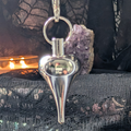 Grey metal teardrop shaped pendulum with black background, soft candlelight and amethyst geode
