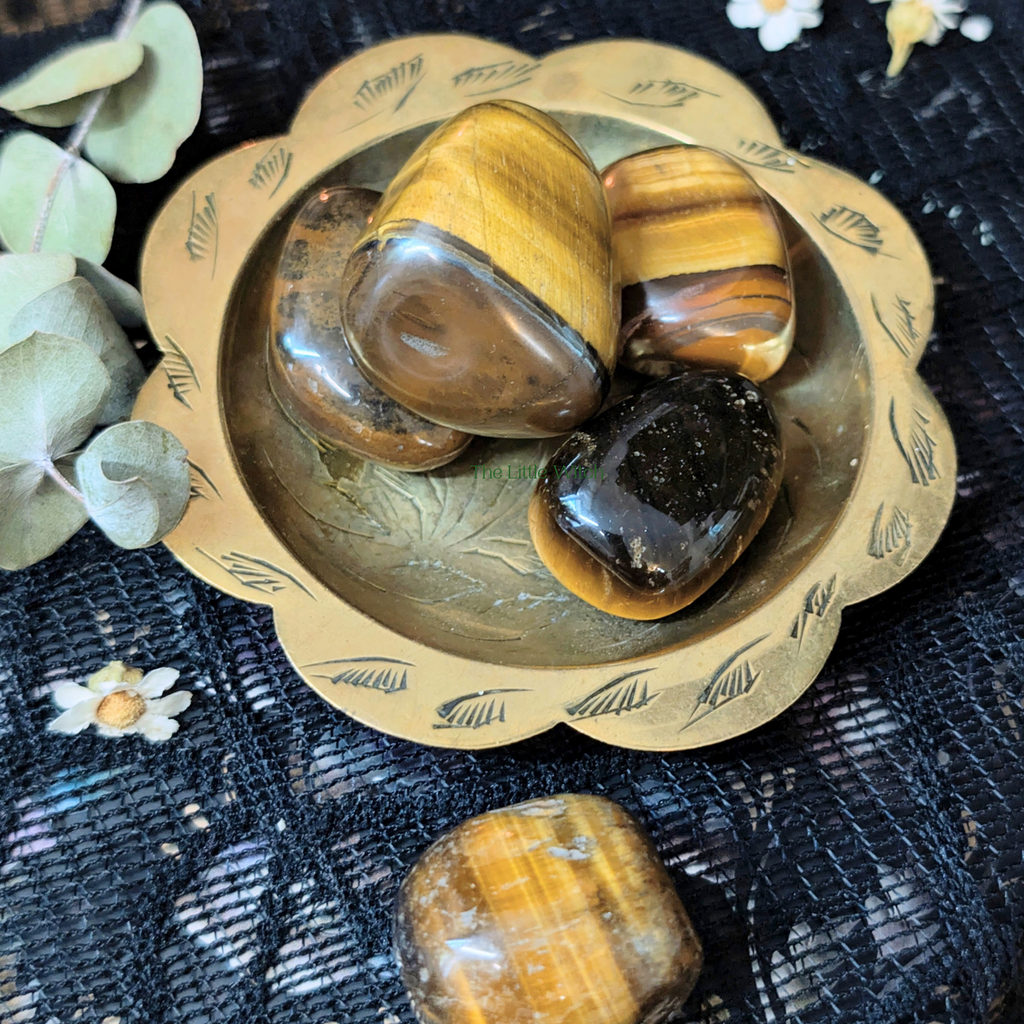Three tiger's eye stones in a decorative brass bowl on a dark textured surface with eucalyptus leaves.