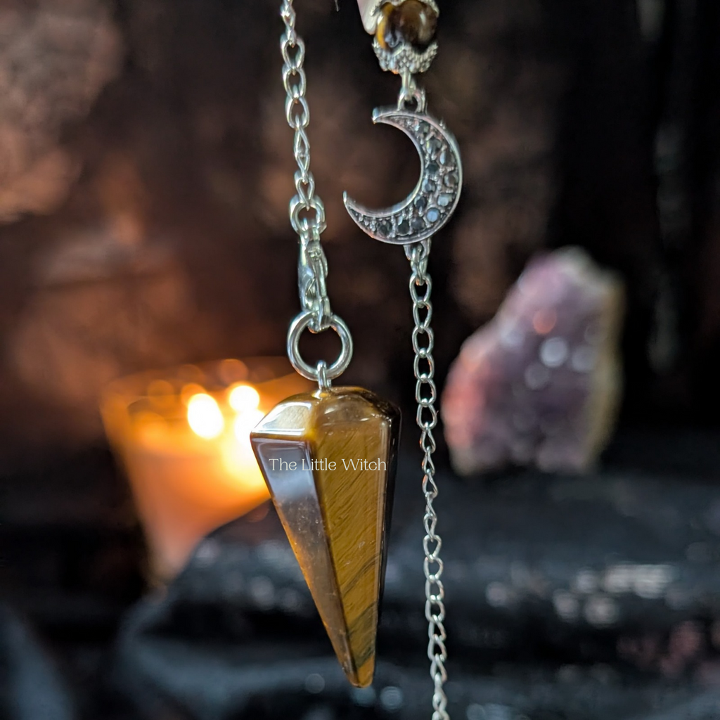 Tiger eye pendulum with moon charm on a chain against a mystical background with candles and crystals.