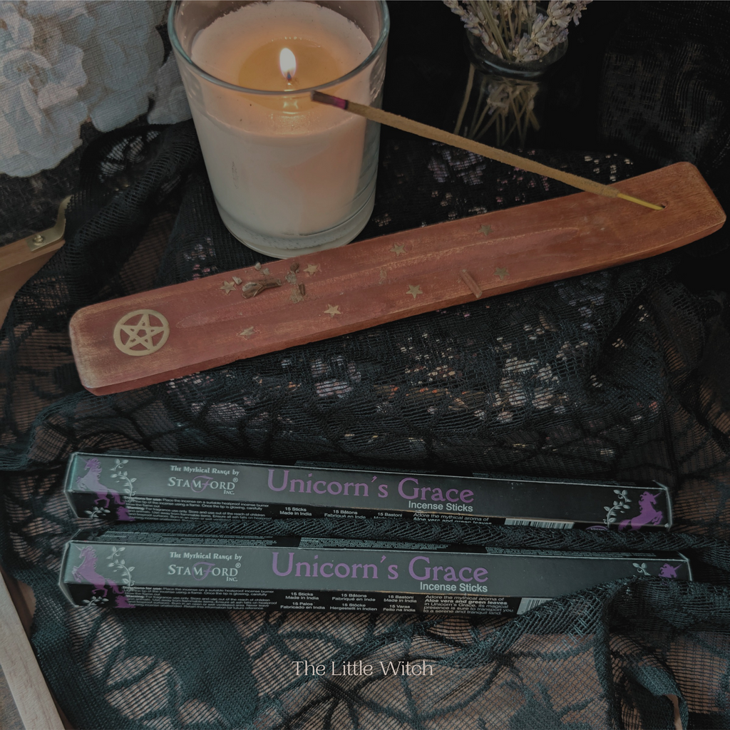 Unicorn's Grace incense sticks with a lit candle and pentacle symbol on a dark fabric background.