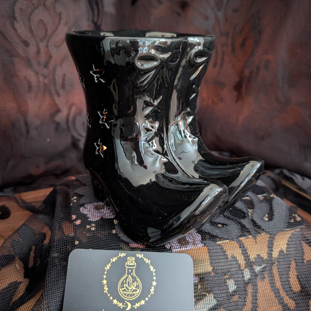 A glossy black ceramic boot-shaped wax warmer with small cutout details, displayed on a patterned fabric surface next to a dark card with a gold illustration.