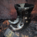 A glossy black ceramic boot-shaped oil burner with star-shaped cutouts, displayed on a black lace fabric surface with a branded card in the foreground.