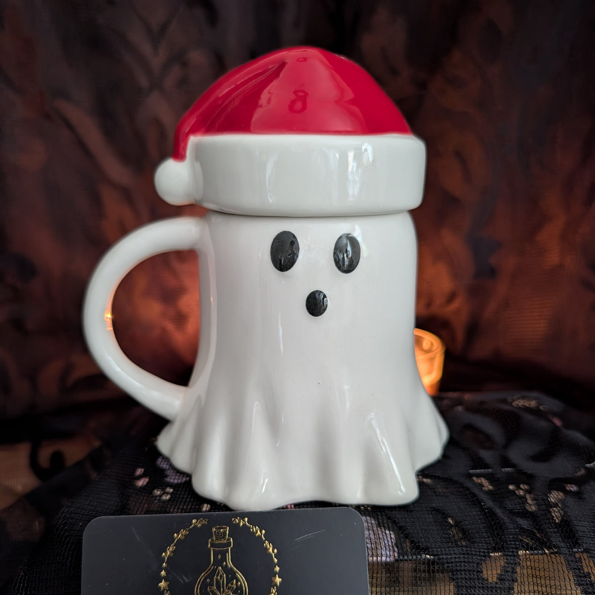 A white ceramic ghost-shaped mug with a red Santa hat lid, featuring black eyes and a black mouth, set against a dark, moody background.