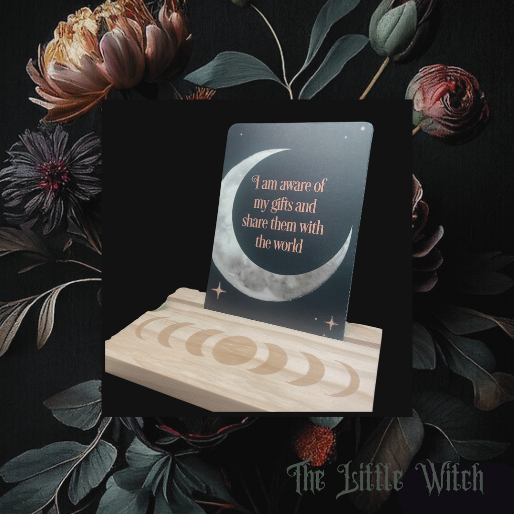 A set of Affirmation Cards displayed on a wooden stand with floral background. The card in the foreground features text and a crescent moon design.