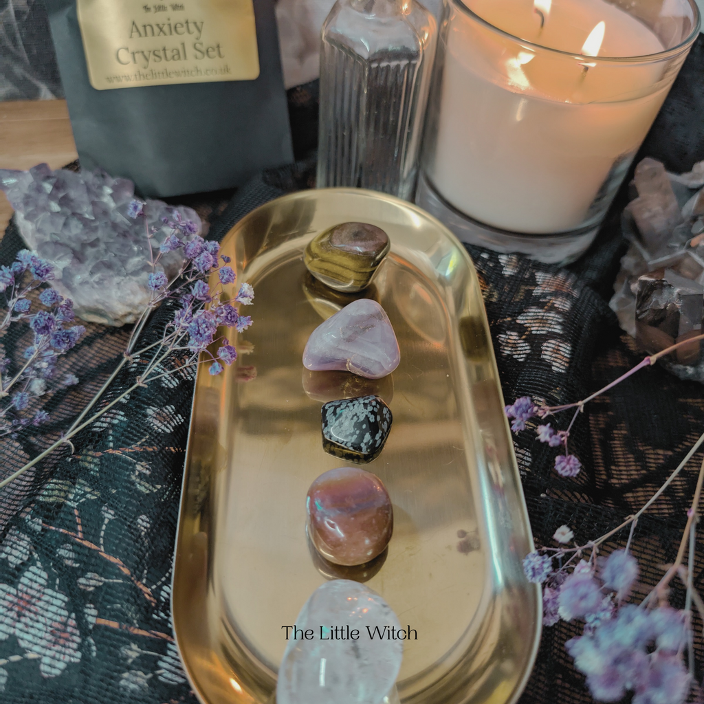 An Anxiety Crystal Set displayed on a gold tray with a candle and purple flowers in the background, including various tumbled crystals and a black pouch with the brand name 'The Little Witch' visible.