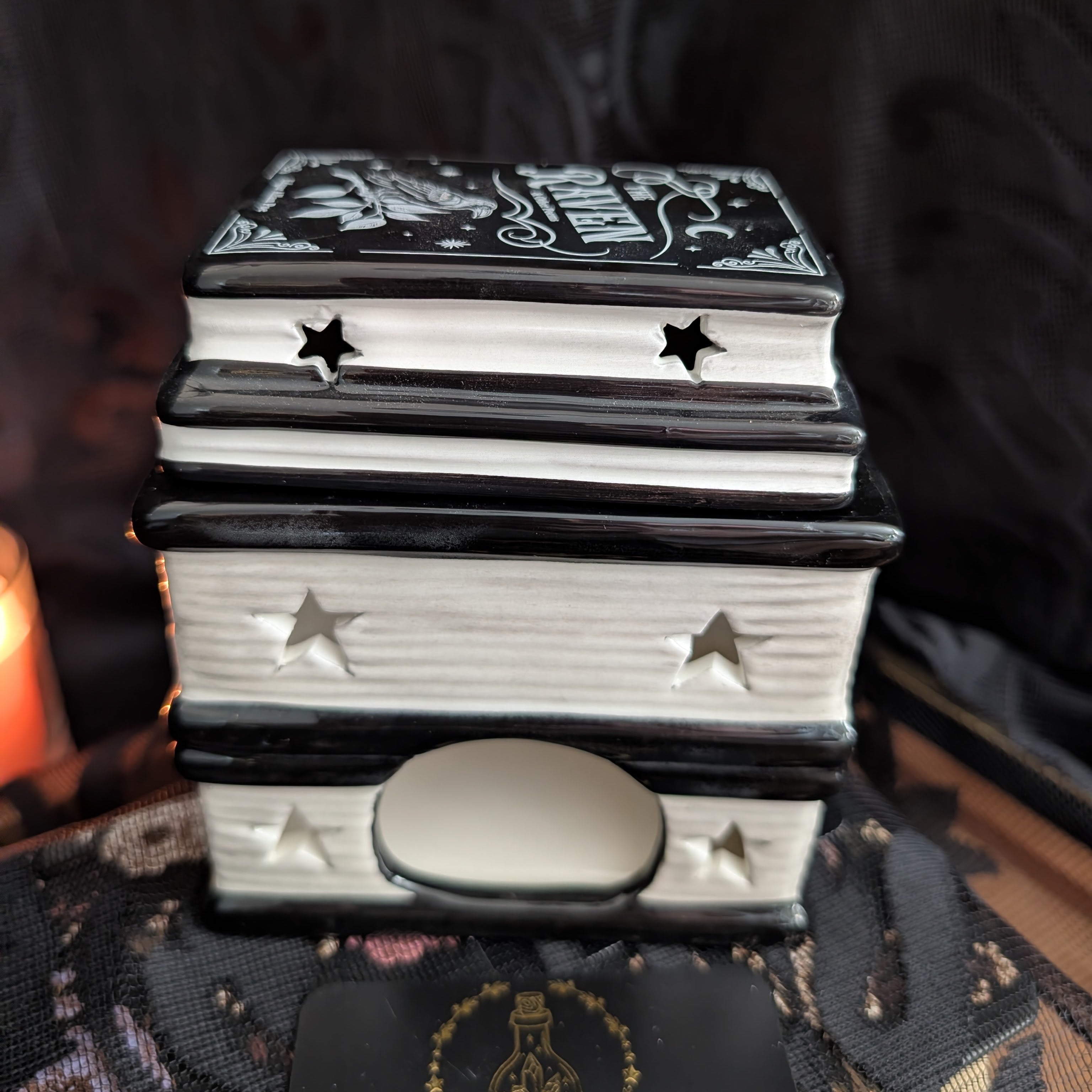 A black and white ceramic book stack box with star cutouts and a small oval drawer front, photographed in a moody setting with a candle and dark fabric in the background.