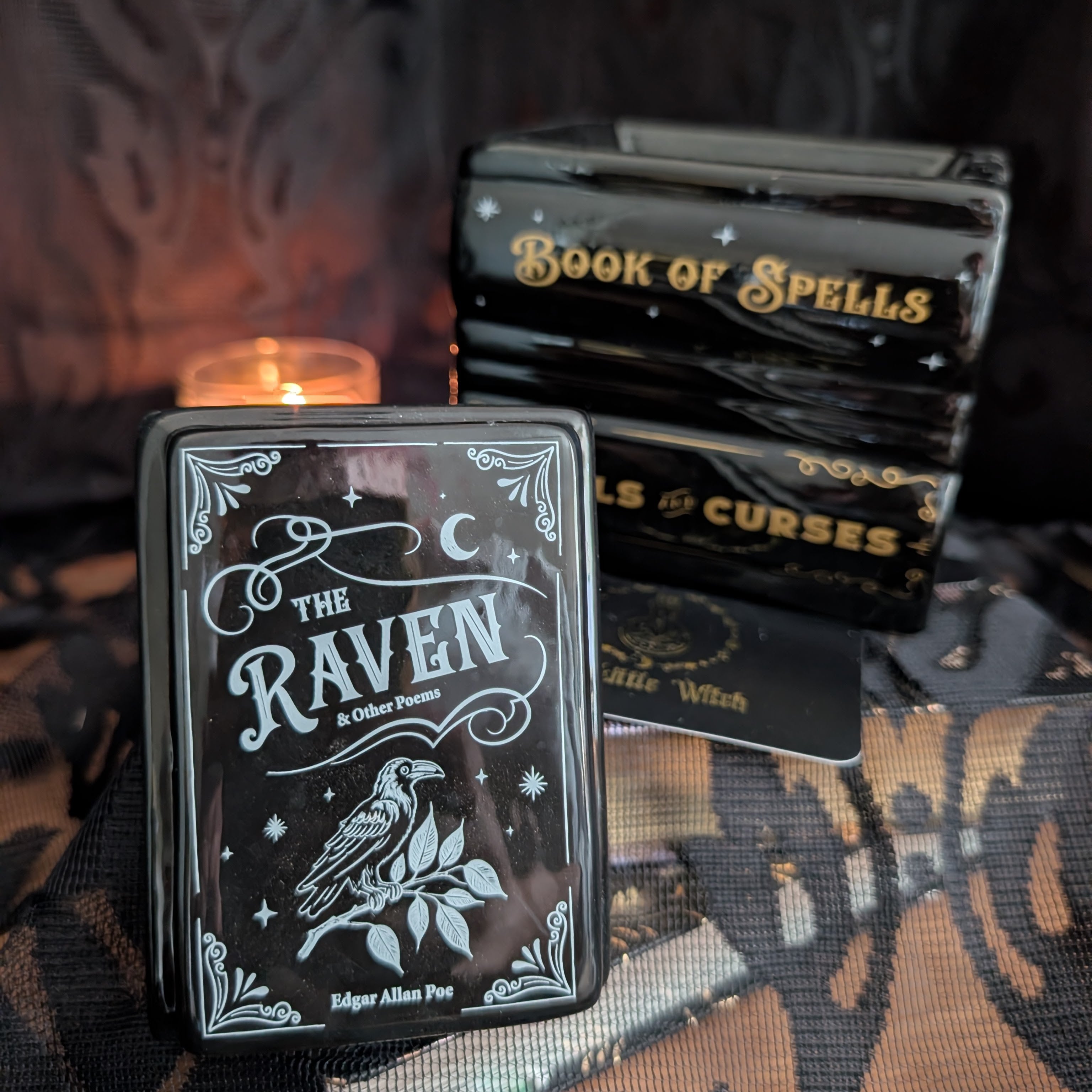 A black book stack  box titled 'The Raven' in Edgar Allan Poe style, shown in front of a stack of matching boxes labeled 'Book of Spells' and 'Curses'. A small lit candle glows in the background against a dark, patterned fabric backdrop.