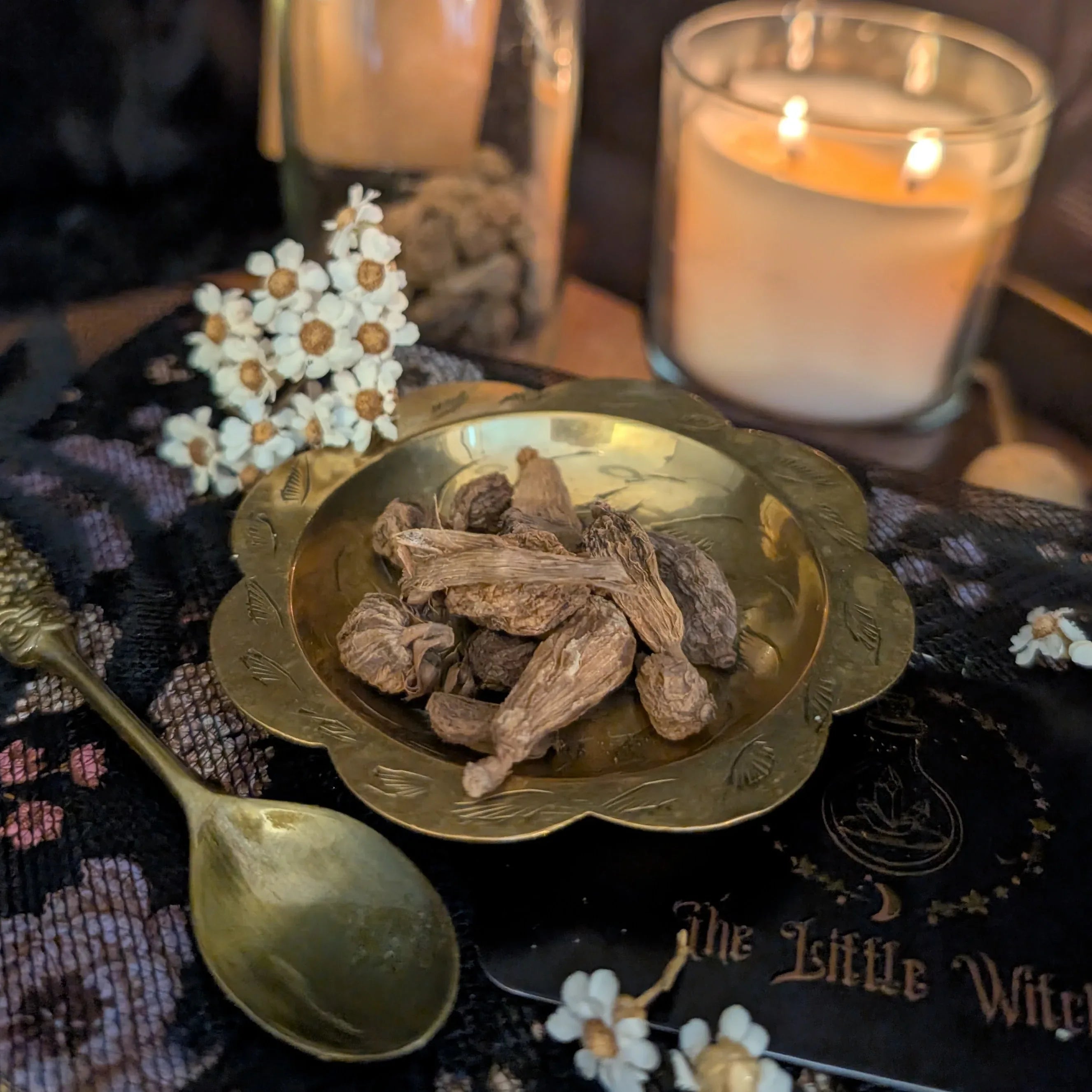 Gold dish with dried roots on a dark fabric background with candles and flowers.