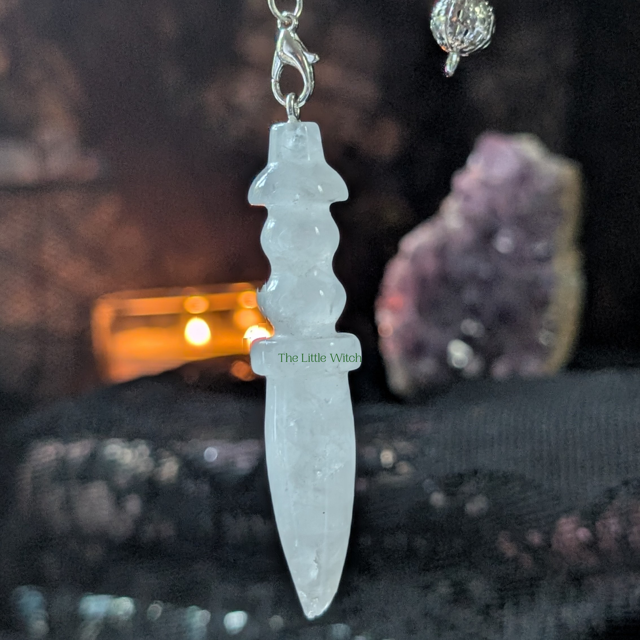 A dark aesthetic of black lace with soft candlelight and an amethyst geode is the backdrop of a clear quartz pendulum 