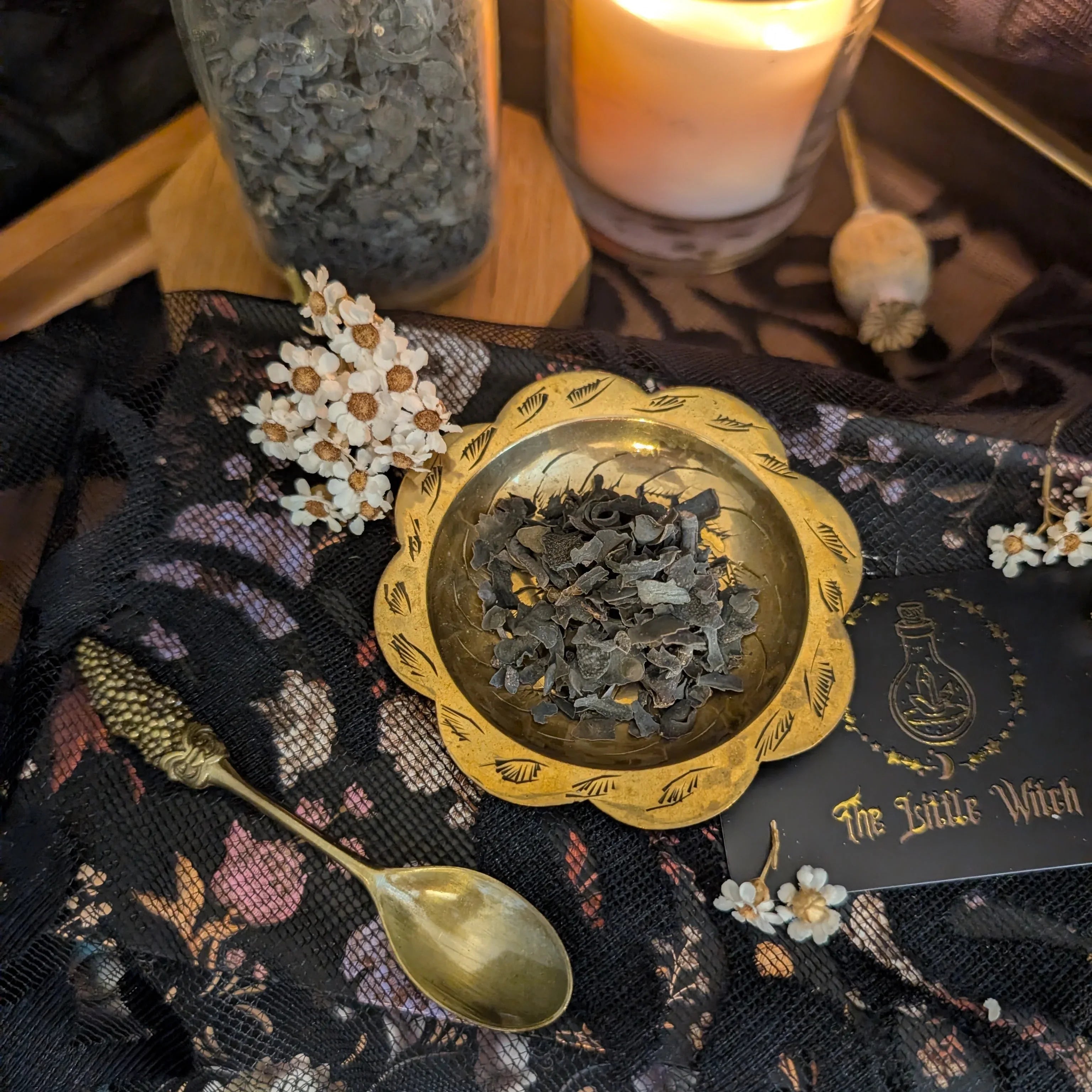 Decorative gold bowl with herbs on a black cloth with floral patterns, surrounded by candles and a spoon.