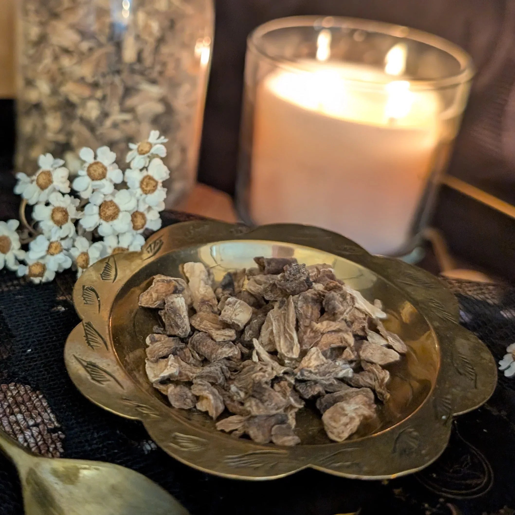Gold bowl with dried herbs on a dark surface with a lit candle in the background