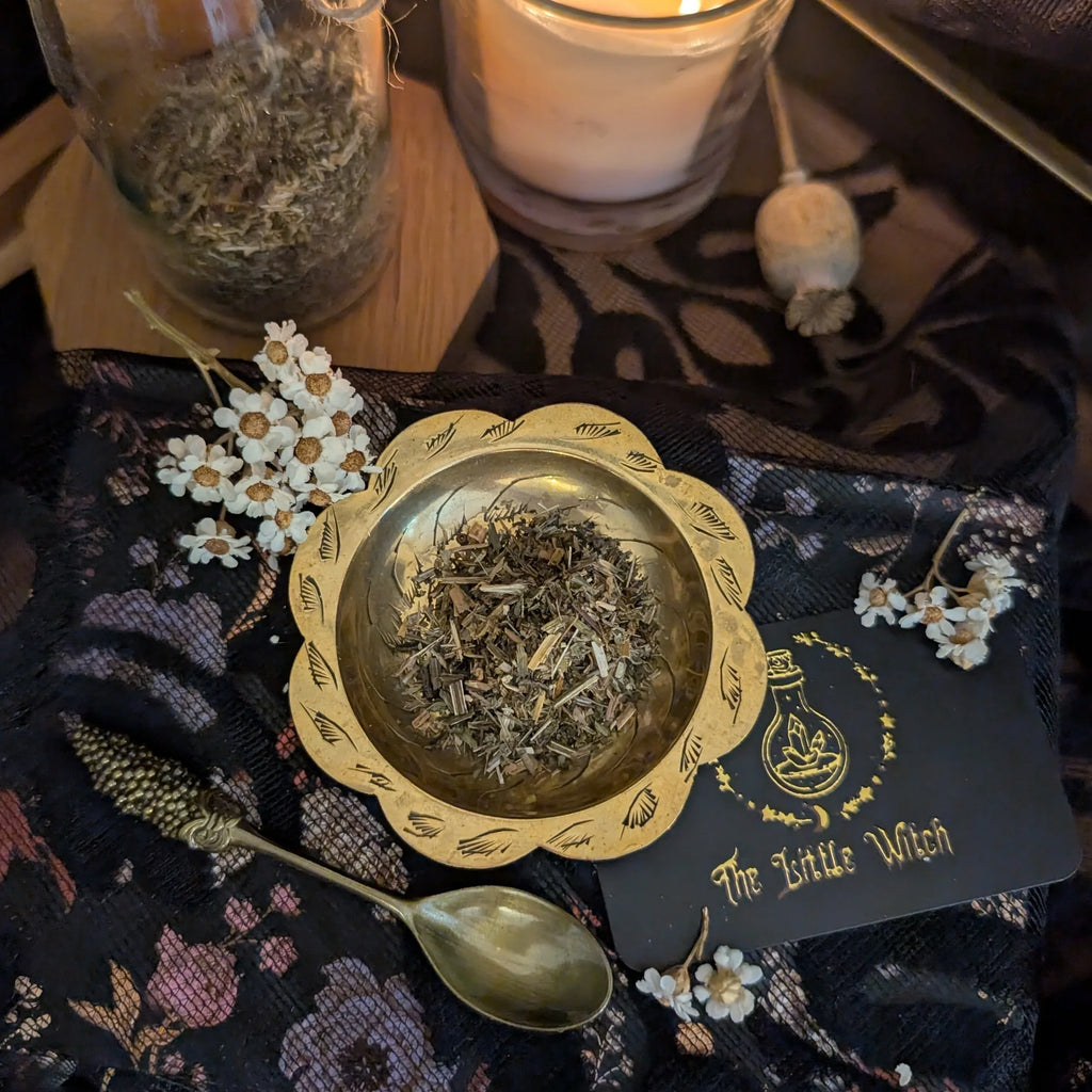 Gold bowl with dried herbs on a dark fabric background with 'The Little Witch' card.